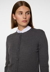 Grå stickad cardigan med rund halsringning och knäppning. Slät struktur, långa ärmar, subtil stygndetaljering runt kanterna.