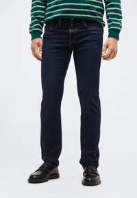 Άνδρας που φορά στενά τζιν σκούρου μπλε, μαύρα δερμάτινα παπούτσια και πράσινο-λευκό ριγέ πουκάμισο με μακριά μανίκια, στέκεται μπροστά σε απλό φόντο.