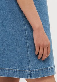 Blaue Jeansrock mit sichtbaren Nähten am Saum. Eine Hand ruht auf dem Rock und betont dessen glatte Textur und die taillierte Form.