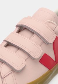 Ανοιχτό ροζ αθλητικό παπούτσι με τρεις ιμάντες βέλκρο και κόκκινο λογότυπο "V" στο πλάι, με υφασμένη μπεζ σόλα.