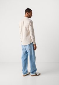Man staat zijwaarts, draagt een beige langemouwen shirt, losse lichtblauwe jeans en witte sneakers met zwarte strepen op een witte achtergrond.