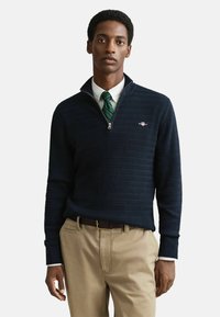 Navy pullover trui met een hoge kraag en een kwartrits, met een textuurpatroon en een klein logo aan de linkerkant. Gemaakt met kaki broek.