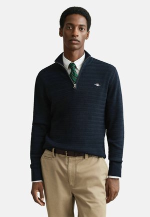 Marineblauer Pullover mit hohem Kragen und einem Viertelreißverschluss, der ein strukturiertes Muster und ein kleines Logo auf der linken Seite trägt. Kombiniert mit khakifarbenen Hosen.