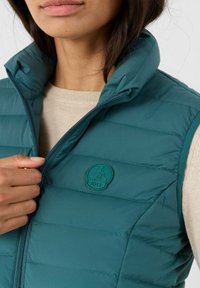 Gilet imbottito color turchese con colletto alto, cuciture orizzontali e un patch logo circolare. Chiusura con zip frontale e strato interno morbido visibile.