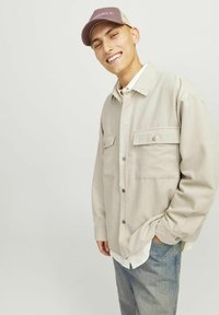 Light beige cord jacket with two chest pockets, button front, and ribbed texture, paired with light blue jeans and a pink cap.