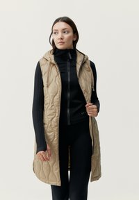 Beige vadderad väst med huva, med en dragkedja fram, fickor och dragsnören; bärs över en svart långärmad topp ochleggings.