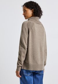 Beige gebreide trui met een hoge kraag, geribde zoom en gestructureerde schouderaccenten. Gepaard met blauwe denim jeans.