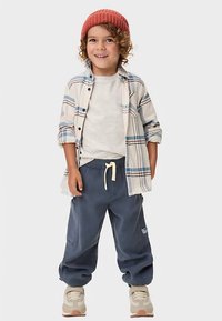 Dítě v kostkované košili zapínané na knoflíky v bílé, modré a béžové barvě přes světle barevné tričko, doplněné tmavě modrými tepláky a béžovými teniskami.