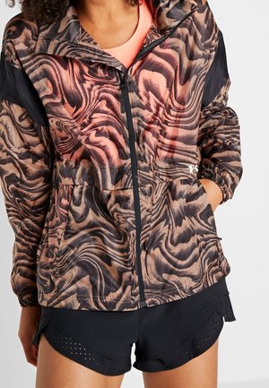 Chaqueta ligera con un patrón ondulante en marrón y rosa, con cremallera fronta, puños elásticos y detalles en negro. Llevada sobre pantalones cortos deportivos negros.