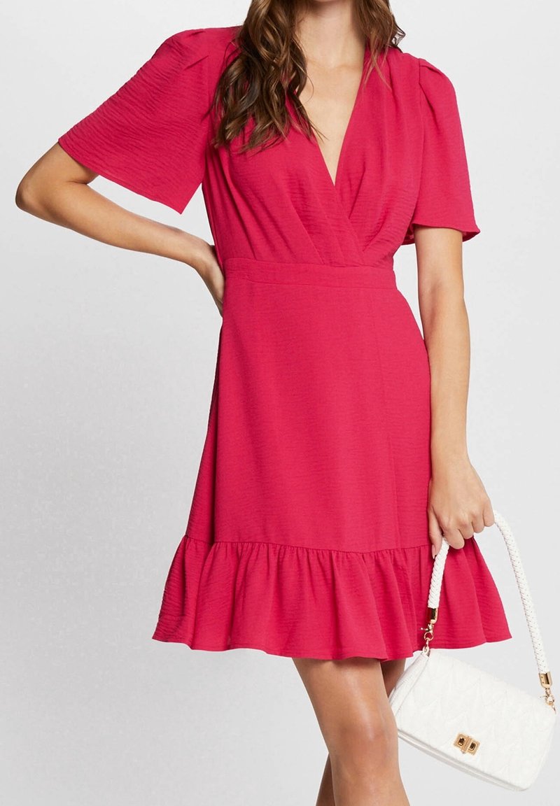 Femme portant une robe rose à manches courtes avec un ourlet à volants, tenant un sac à main blanc matelassé avec des ferrures dorées.