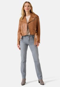 Braune Leder-Cropped-Jacke mit Druckknopf- und Reißverschlussdetails, kombiniert mit hellgrauen Jeans und grauen, figurschmeichelnden Ankle Boots.
