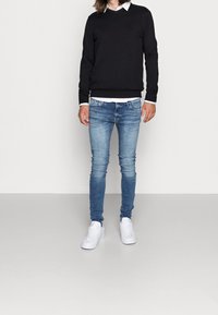 Maglione nero all'uncinetto, camicia bianca con colletto sotto, jeans skinny blu chiaro e sneakers bianche. Tessuti morbidi e vestibilità casual.