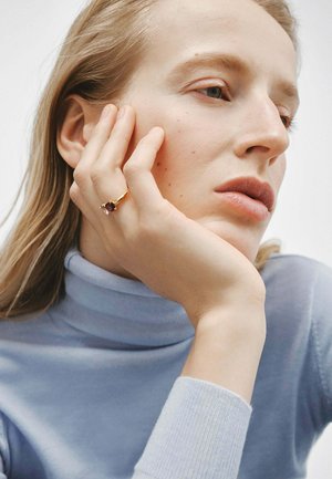 Anillo de oro con una piedra preciosa púrpura, llevado en una mano contra un cuello de tortuga azul pálido. La textura suave complementa la piel lisa de la modelo.