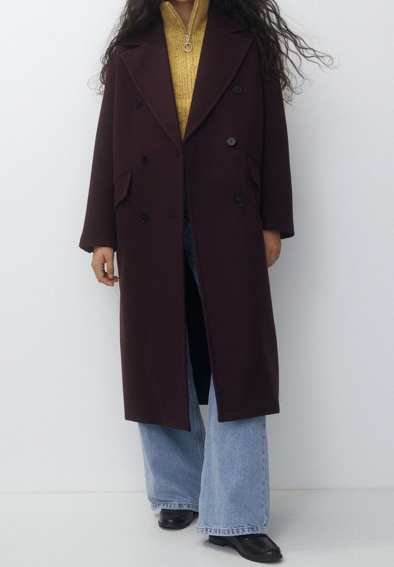 Femme portant un long manteau marron foncé croisé sur un pull zippé jaune moutarde, un jean large bleu clair et des chaussures noires.