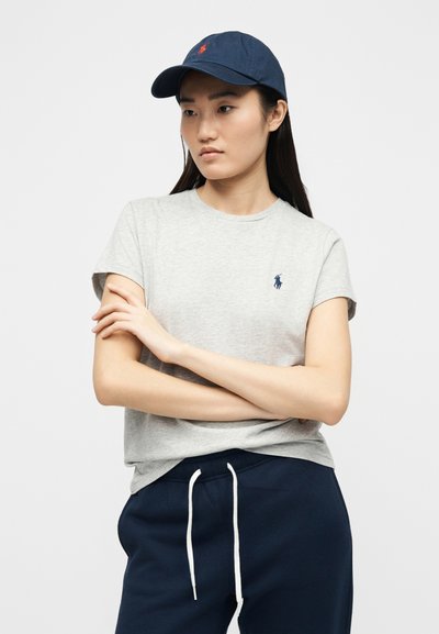 Γυναίκα που φορά γκρι μπλουζάκι με λογότυπο σε ναυτικό μπλε, σκούφο ναυτικού μπλε με κόκκινο λογότυπο και ναυτικό μπλε παντελόνι με λευκά κορδόνια, όρθια με σταυρωμένα χέρια.