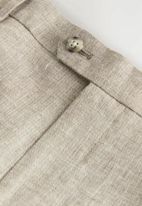 Vita di pantaloni in tessuto beige testurizzato con un singolo bottone rotondo e passanti per cintura visibili su sfondo semplice.