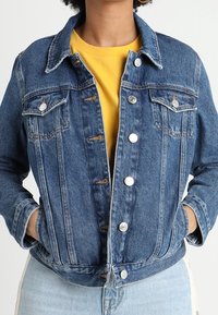 Dunkelblaue Jeansjacke mit durchgängigem Knopfverschluss, zwei Brusttaschen und taillierter Passform. Getragen über einem gelben Hemd mit hochgekrempelten Ärmeln.