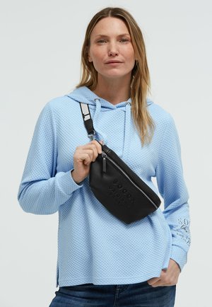 Frau in hellblauer, strukturierter Kapuzenjacke hält eine schwarze Umhängetasche mit Reißverschluss und verstellbarem Riemen vor einem einfarbigen Hintergrund.