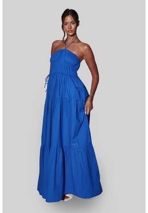 Kobieta w niebieskiej, warstwowej sukni maxi bez rękawów z wiązaniem na szyi, stojąca z jedną ręką trzymającą suknię, na szarym tle.