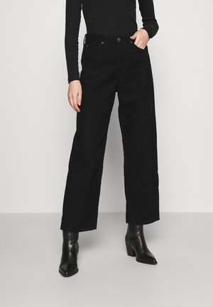 Persoon die zwarte, wijde broek van corduroy draagt, een zwarte top met lange mouwen en zwarte enkellaarsjes met hak, staand tegen een effen achtergrond.