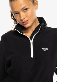 Black fleece half-zip pullover with white zipper and heart logo patch. Soft texture, relaxed fit, standing collar.