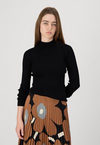 Fekete bordás garbós pulóver hosszú ujjal, karcsú fazonnal. A modell egy pliszírozott, mintás barna szoknyával viseli.