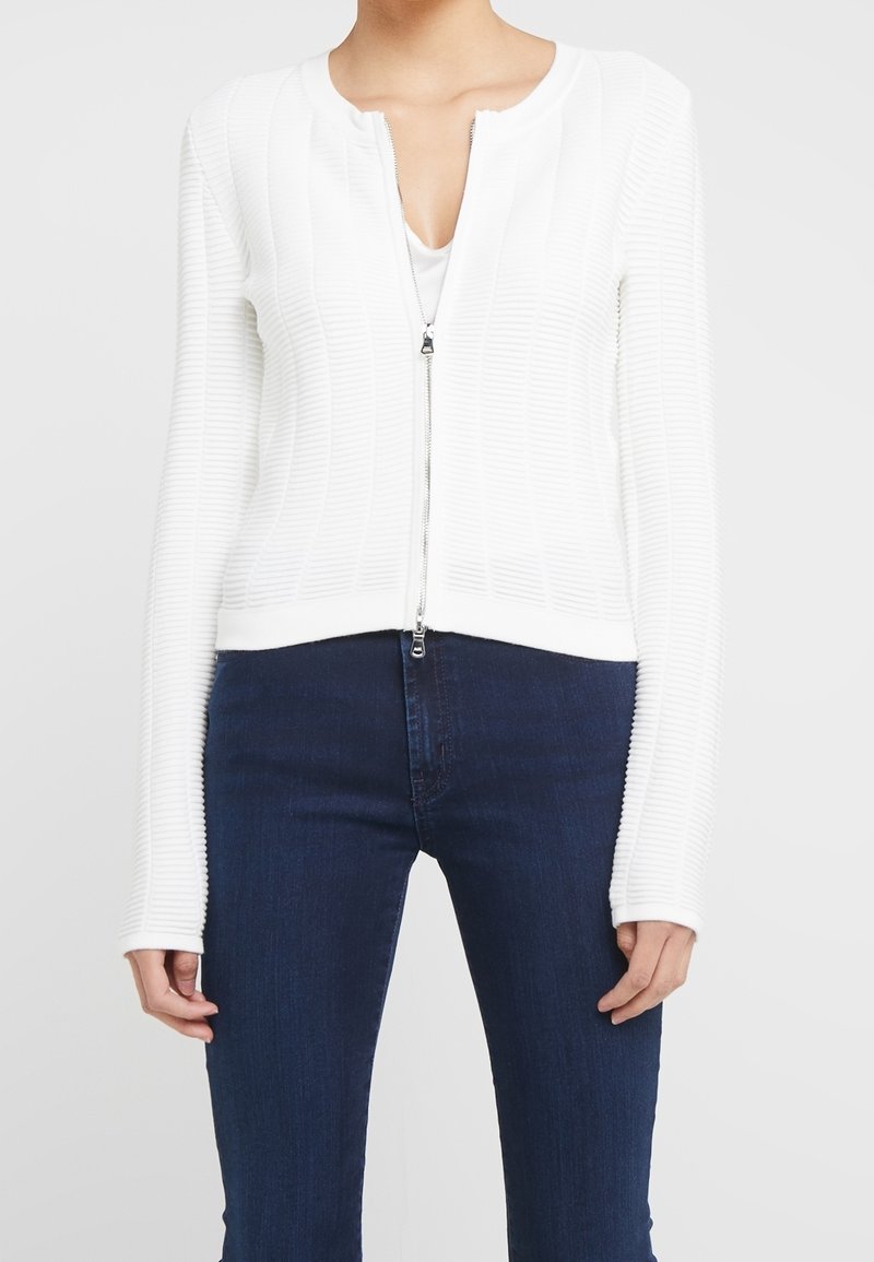 Biały kardigan na zamek błyskawiczny z prążkowaną teksturą, długimi rękawami i krótko skrojonym fasonem. W zestawieniu z ciemnoniebieskimi dżinsami dla kontrastu.