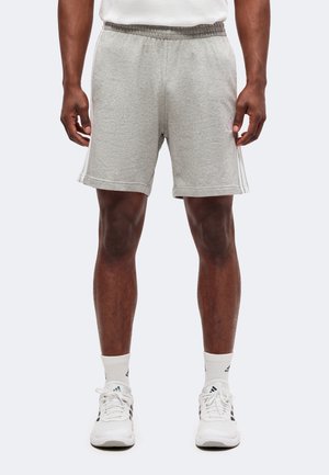 Mężczyzna w jasnoszortach sportowych, białych butach Adidas i białych skarpetach, stojący ze swobodnie opuszczonymi rękami.