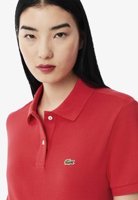 Röd polo skjorta i texturerad tyg, med en treknappsknäppning och en liten grön krokodil-logotyp på vänster bröst.