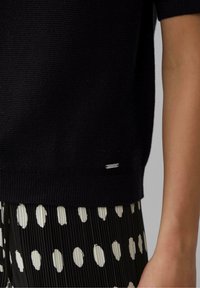 Suéter negro de punto con bordes acanalados, que presenta una pequeña etiqueta metálica. Combinado con una falda plisada a rayas blancas y negras con patrones ovalados.
