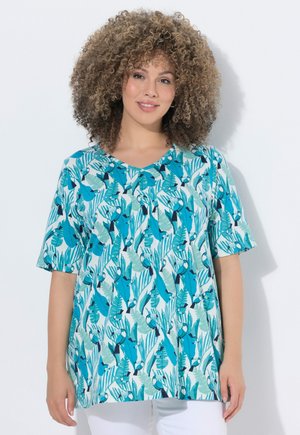 Kobieta z kręconymi włosami, ubrana w krótkim rękawem, niebiesko-zielony tropikalny top i białe spodnie, stoi na białym jednolitym tle.