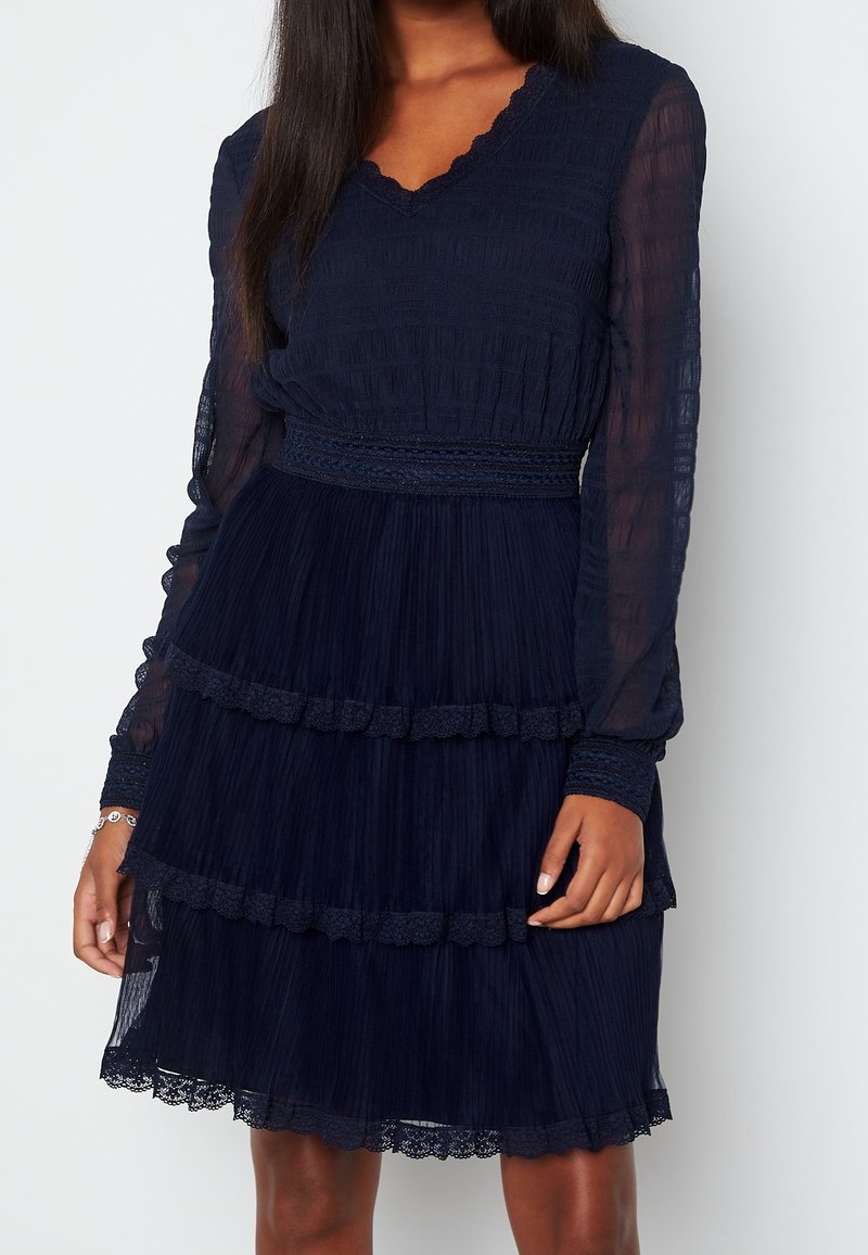 Robe bleu marine à manches longues et transparentes, design plissé, volants superposés à l'ourlet et accents en dentelle festonnée au col et aux poignets.