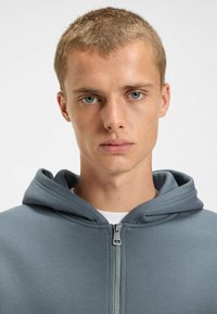 Grijze zip-up hoodie van een gestructureerde stof, met een strakke capuchon en een gladde rits. Draagt over een wit shirt, minimalistisch ontwerp.