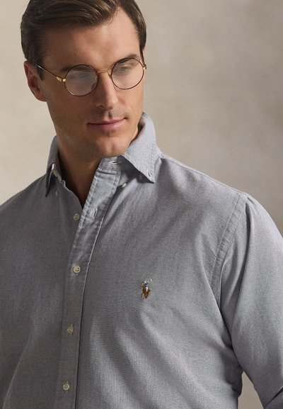 Homme portant des lunettes à monture dorée et une chemise gris clair boutonnée avec un petit logo brodé de joueur de polo sur la poitrine gauche.