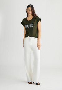 T-shirt verde oliva a maniche corte con logo "LIU-JO" in paillettes, abbinata a pantaloni bianchi a gamba larga e sandali neri con punta aperta.