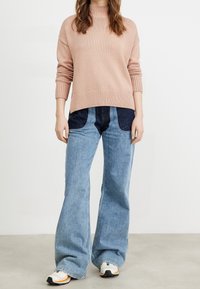 Hellrosa gestrickter Pullover mit gerippten Bündchen, kombiniert mit hellblauen Schlagjeans aus Denim, die dunkle Denim-Patches an der Taille haben. Weiße Sneakers.