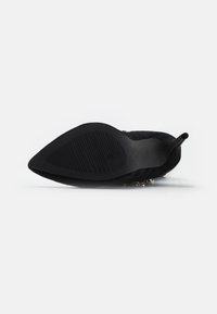 Zapato de tacón alto de ante negro con punta afilada, suela texturizada y adornos decorativos a lo largo de los bordes.