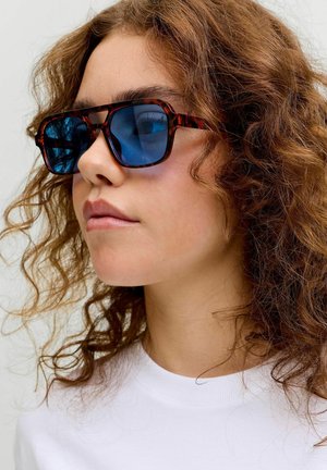 Jonge persoon met krullend roodbruin haar, die een zonnebril met schildpadmontuur en blauwe glazen draagt en een wit shirt met ronde hals aanheeft.