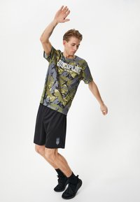 Conjunto deportivo que incluye una camiseta de malla en camuflaje verde y gris con el texto "LA DISCIPLINA ES LA CLAVE" y pantalones cortos negros con detalles grises.
