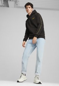Felpa nera con strisce laterali gialle e logo, abbinata a jeans azzurri chiari e sneaker bianche con dettagli neri.