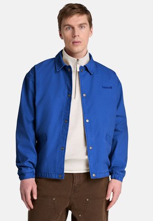 Ung mand iført en skarp blå knapjakke over en hvid lynlås sweater og brune bukser, stående mod en ensfarvet baggrund.
