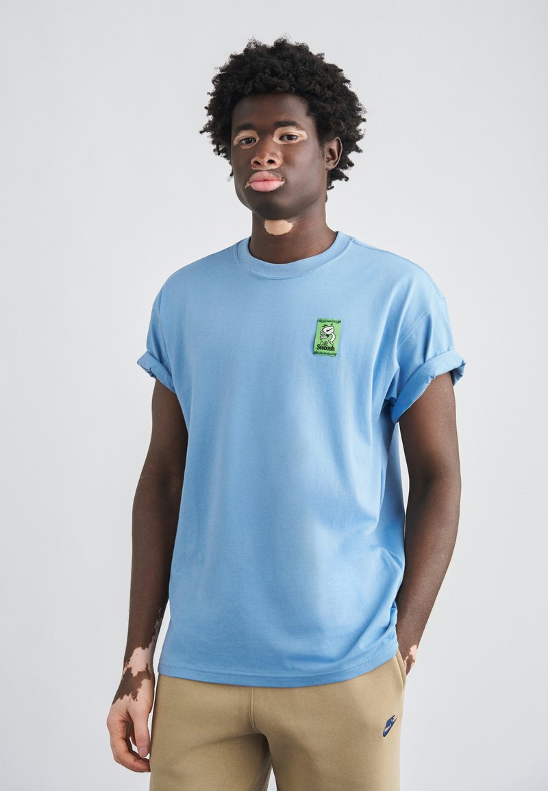 Gaiši zila kokvilnas T-krekls ar uzlocītām piedurknēm, kreisajā krūtīs zaļa izšūta uzlīme ar karikatūras dizainu. Saskaņots ar bēšām biksēm.