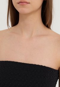 Melna strapless blūze ar teksturētu virsmu, kurai ir ģeometrisks raksts gar kakla izgriezumu un gluda auduma. Nav redzamu elementu vai akcentu.