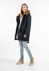 Parka blu scuro con bordo in pelliccia sintetica, texture liscia, lunghezza media, due tasche anteriori, indossato sopra una maglietta bianca e jeans strappati blu chiaro.