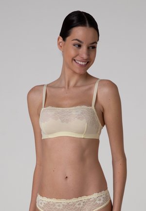 Donna con capelli scuri con reggiseno a fascia in pizzo crema e mutandine di pizzo abbinate, che sorride e guarda di lato su uno sfondo semplice.