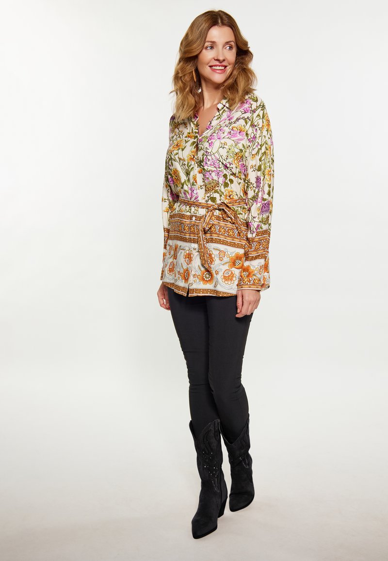 Blusa a fiori in crema con fiori viola, gialli e arancioni; vita cintata; abbinata a pantaloni skinny neri e stivali neri.