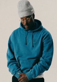 Mężczyzna w szarej dzianinowej czapce, okularach z złotymi oprawkami i niebieskiej bluzie, spoglądający w dół z dłońmi splecionymi przed sobą na jednolitym tle.