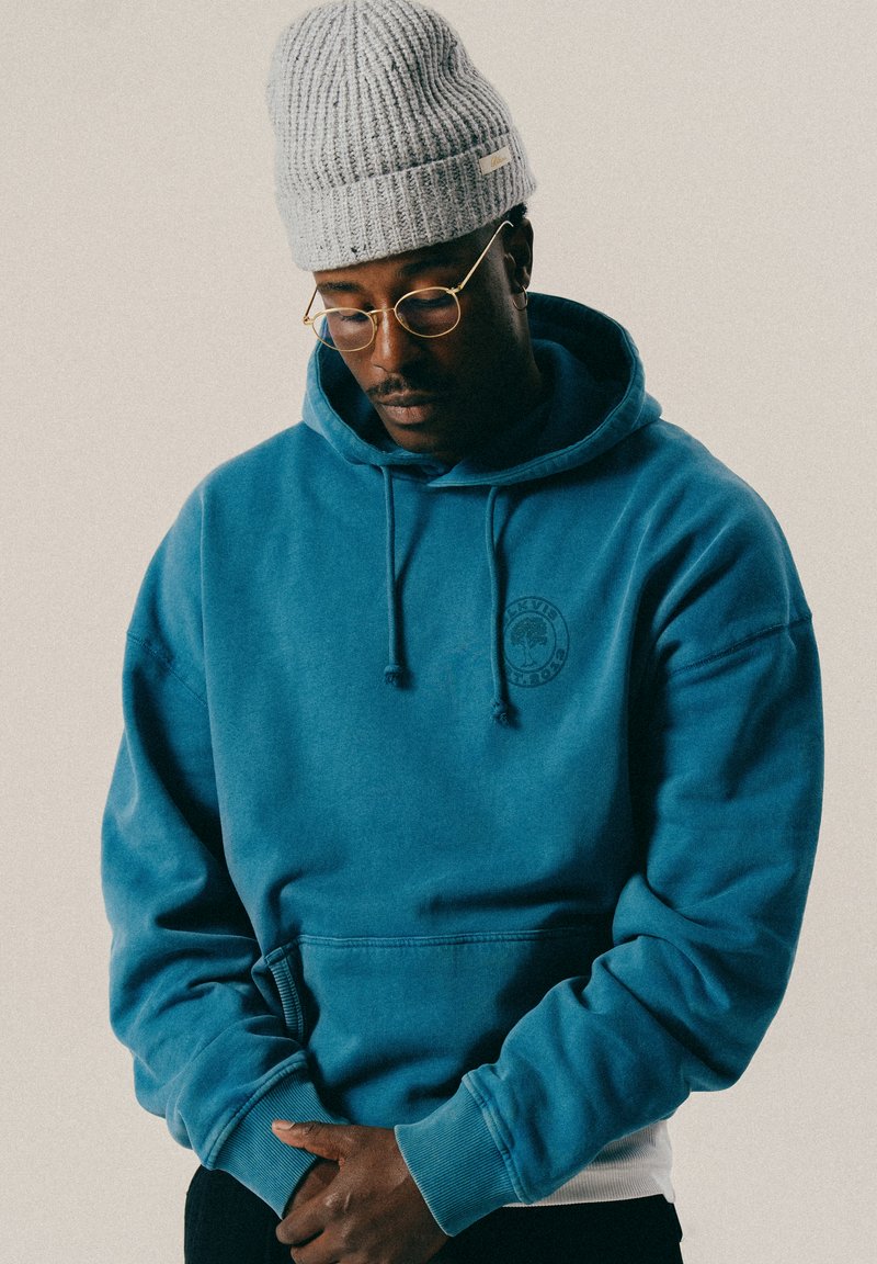 Mężczyzna w szarej dzianinowej czapce, okularach z złotymi oprawkami i niebieskiej bluzie, spoglądający w dół z dłońmi splecionymi przed sobą na jednolitym tle.