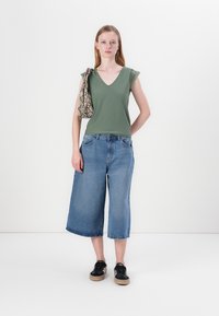 Grønn v-hals topp med blonde detaljer, blå vide denim culotter og svarte sneakers. Modellen holder en mønstret håndveske.