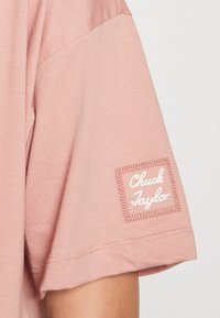 Světle růžové bavlněné tričko s kulatým výstřihem a krátkými rukávy. Obsahuje přišitou obdélníkovou etiketu s nápisem "Chuck Taylor" v bílé barvě.